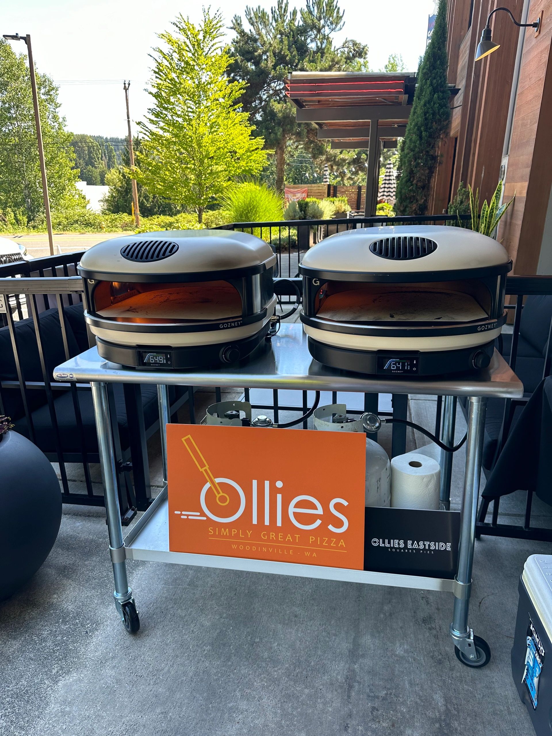 Two portable stone-deck pizza ovens on a stainless cart with the orange Ollies signage, set up at an event in Woodinville.